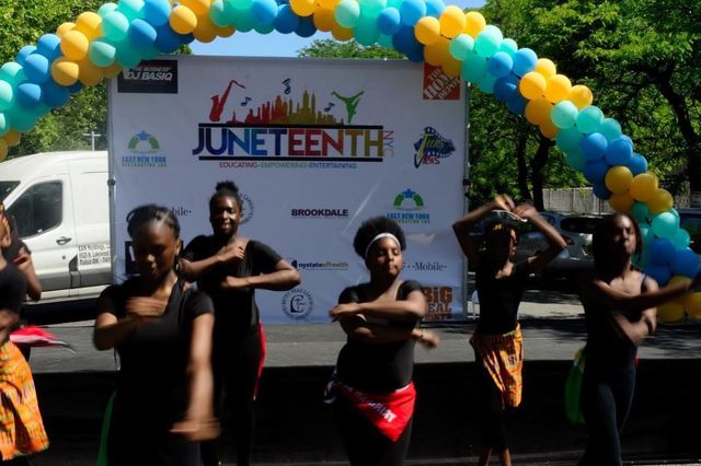 A group of young Black women perform in black outfits in front of a Juneteenth NYC sign